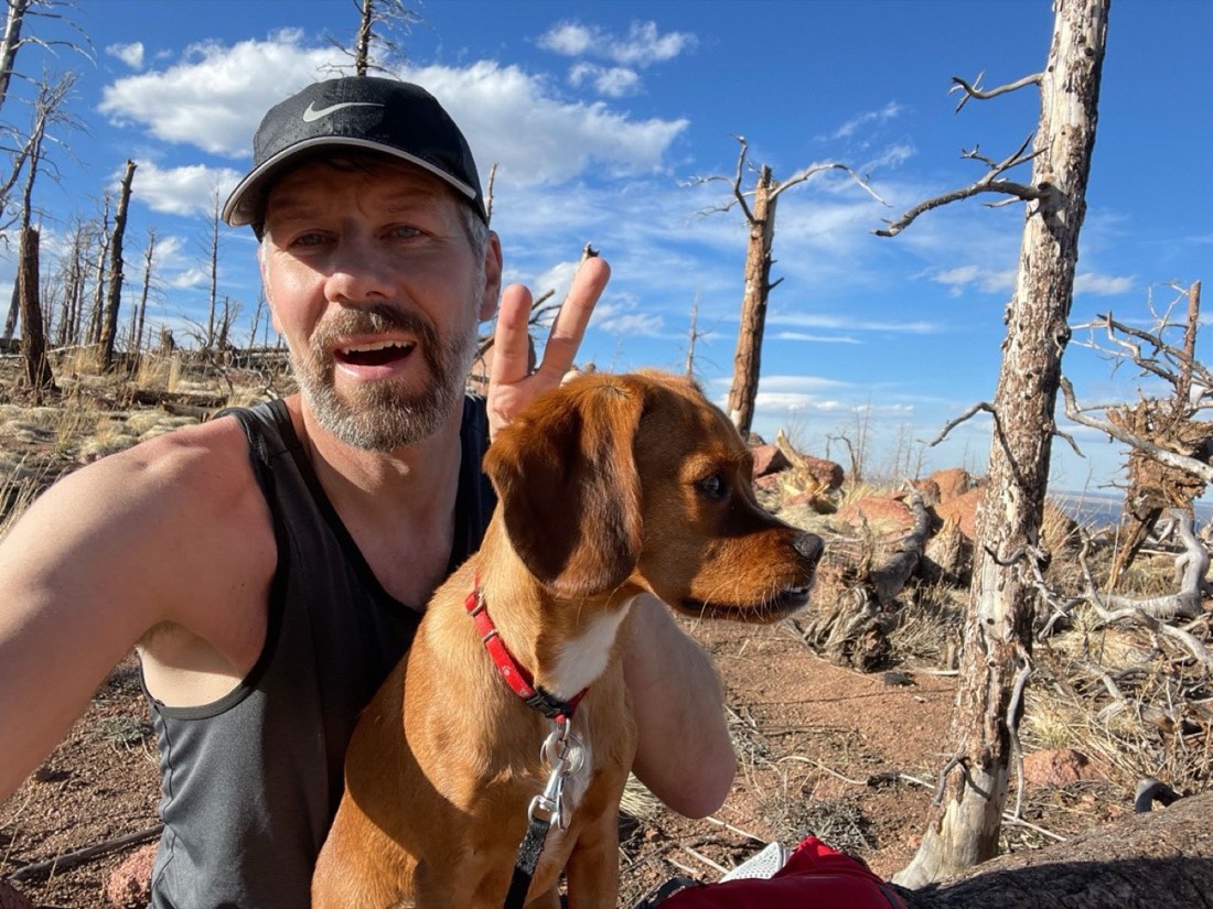 Brian in a running cap flashing a peace sign beside his dog on a burned-forest trail under a blue Colorado sky