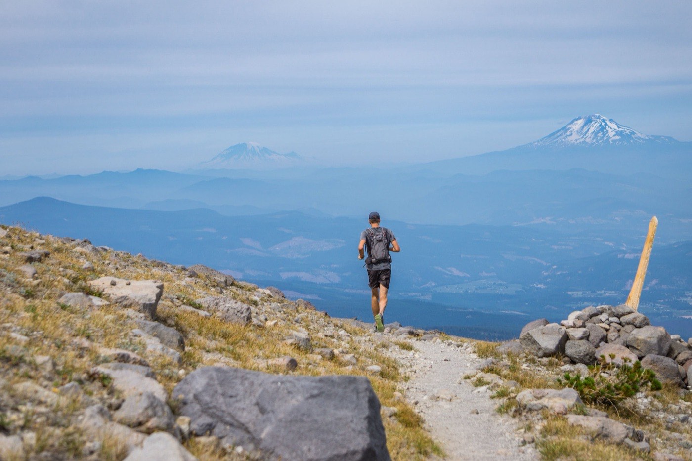 Trail runner descending a rocky high-alpine trail, hazy blue ranges and two snow-capped volcanoes on the horizon